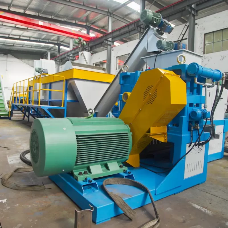 a comprehensive view of an industrial plastic recycling facility, featuring a variety of specialized equipment. This setup includes a series of machines such as conveyors, shredders, and separators, all crucial for the recycling process. The machinery is predominantly painted in vibrant blue and yellow, colors often used to denote machinery and caution in industrial environments. This particular section of the facility appears to focus on the initial stages of recycling where plastics are sorted, cleaned, and prepared for further processing. Visible in the scene are large motors and mechanical parts, suggesting these machines handle heavy-duty processing tasks like shredding and crushing. The background shows additional parts of the facility, hinting at a large-scale operation designed to process significant volumes of material efficiently.