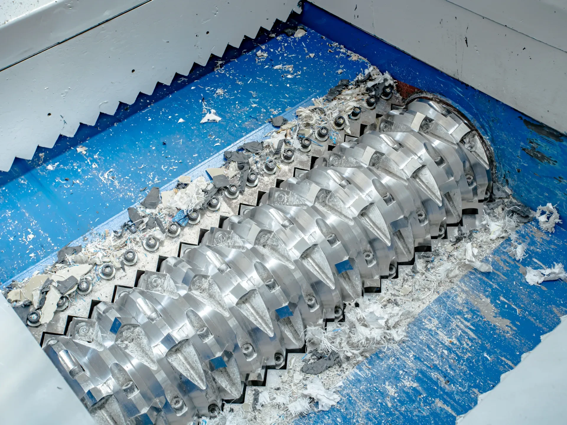 Close-up view of a single-shaft shredder's cutting mechanism. The image shows the robust and sharp cutting blades of the shredder, designed to handle and break down various materials. The blades are mounted on a rotating shaft within a blue and white cutting chamber. Pieces of shredded material are visible, showcasing the shredder's effectiveness in processing waste. This machinery is ideal for recycling and waste processing applications, offering high efficiency and durability.